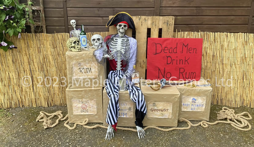 Decorations for entrance to a pirate party - skeleton sits atop boxes of rum and gunpowder with a backdrop of reed fencing and a wooden pallet